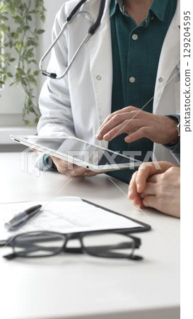 Male doctor in lab coat over a green shirt, and stethoscope gesturing while explaining diagnosis to patient using tablet in modern medical office. Medicine and health care concept 129204595