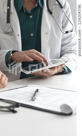 Doctor man in lab coat over a green shirt, and stethoscope is using tablet while consulting patient in modern medical office. Medicine and health care concept Doctor man in lab coat over a green shirt, and stethoscope is using tablet while consulting patient in modern medical office. Medicine and health care concept 129204596