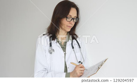 Doctor woman wearing stethoscope, black glasses and white coat over green blouse is standing and writing on a clipboard, representing medical expertise and patient care. Medicine and health care 129204597
