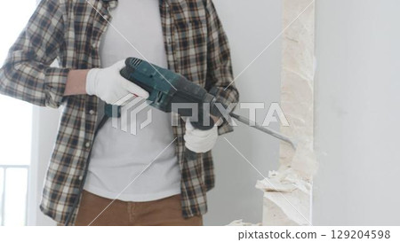 Unrecognizable male construction worker wearing beige checkered shirt and protective gloves, is demolishing white wall with rotary hammer drill, generating dust, close up view. Renovation concept Unrecognizable male construction worker wearing beige checkered shirt and protective gloves, is demolishing white wall with rotary hammer drill, generating dust, close up view. Renovation concept 129204598