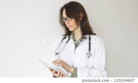Female physician reviewing patient records on digital tablet, standing in contemporary medical workspace with natural lighting. Medicine and health care 129204607