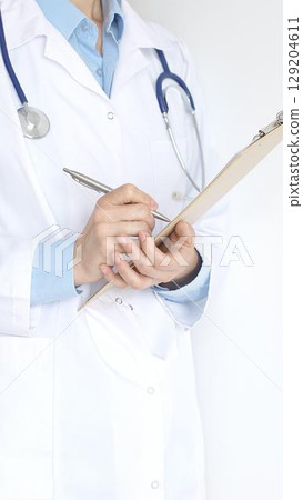 Unknown female doctor wearing stethoscope and white coat over blue blouse is standing and writing on a black clipboard, representing medical expertise and patient care. Medicine concept 129204611