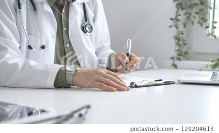 Professional female doctor wearing white lab coat and stethoscope documenting patient information on clipboard near laptop in clinic workspace. Medicine and health care concept 129204613