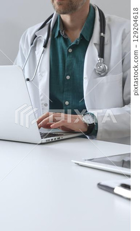Doctor man wearing a white coat over green shirt, and stethoscope is typing on a laptop in a bright medical office, showcasing the intersection of healthcare and technology. Medicine and health care Doctor man wearing a white coat over green shirt, and stethoscope is typing on a laptop in a bright medical office, showcasing the intersection of healthcare and technology. Medicine and health care 129204618