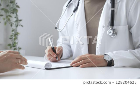 Doctor man listening to patient health concerns and writing a prescription on a clipboard during a consultation in a bright, modern clinic. Medicine and health care concept Doctor man listening to patient health concerns and writing a prescription on a clipboard during a consultation in a bright, modern clinic. Medicine and health care concept 129204623