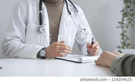 Doctor man is explaining diagnosis to female patient during a medical consultation in a medical office, gesturing with hands over clipboard and white desk. Medicine and health care concept Doctor man is explaining diagnosis to female patient during a medical consultation in a medical office, gesturing with hands over clipboard and white desk. Medicine and health care concept 129204625