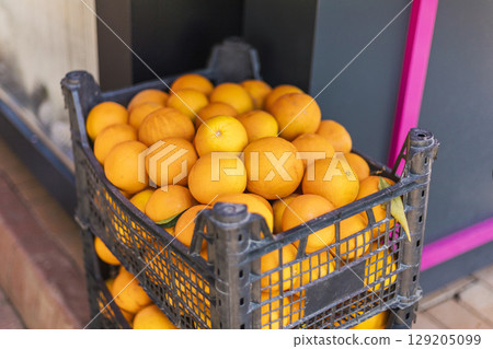 A black crate overflowing with vibrant oranges sits outside a lively market. The sun casts a warm glow on the fruit, highlighting their bright color as shoppers pass by A black crate overflowing with vibrant oranges sits outside a lively market. The sun casts a warm glow on the fruit, highlighting their bright color as shoppers pass by 129205099