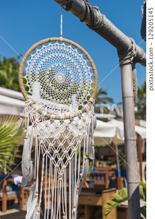 A beautifully crafted dream catcher is suspended from a wooden structure at a tropical restaurant. The intricate design stands out against the bright blue sky, creating a serene atmosphere 129205145
