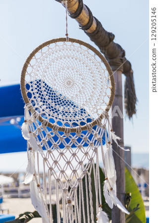 A delicate dream catcher with intricate designs and feathers is suspended from a wooden post at a beach. Bright blue umbrellas enhance the lively atmosphere of the setting A delicate dream catcher with intricate designs and feathers is suspended from a wooden post at a beach. Bright blue umbrellas enhance the lively atmosphere of the setting 129205146