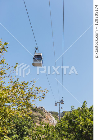 A series of cable cars glide through a clear blue sky, showcasing a beautiful view of lush greenery and rocky hills in the background A series of cable cars glide through a clear blue sky, showcasing a beautiful view of lush greenery and rocky hills in the background 129205174