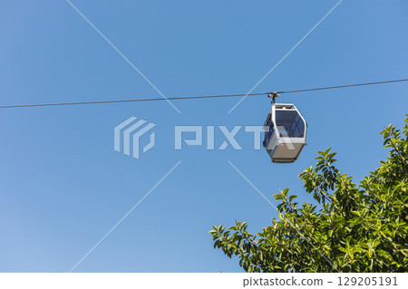 A gondola smoothly travels along its cable, suspended high above lush green trees. The vibrant blue sky adds to the serene atmosphere of this urban landscape A gondola smoothly travels along its cable, suspended high above lush green trees. The vibrant blue sky adds to the serene atmosphere of this urban landscape 129205191