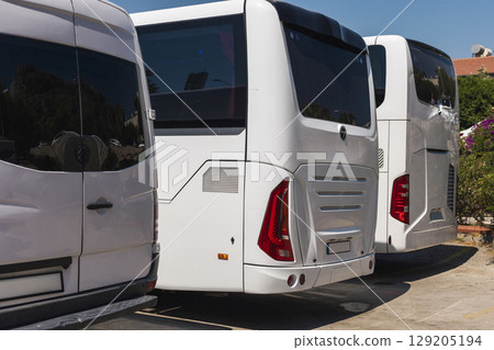 Several white buses are parked closely together at a transportation hub under clear blue skies. The vehicles are in pristine condition, showcasing their sleek designs 129205194