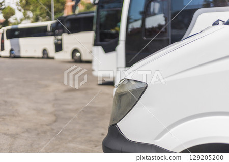 A white van is parked prominently in the foreground of a busy bus terminal filled with several parked buses. Bright daylight highlights the activity and organization in the area A white van is parked prominently in the foreground of a busy bus terminal filled with several parked buses. Bright daylight highlights the activity and organization in the area 129205200