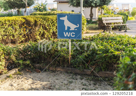 A blue sign featuring a dog symbol indicates a designated restroom area for pets in a park. Surrounding greenery and pathways enhance the outdoor setting for dog owners 129205218