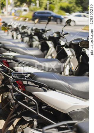 Several parked scooters line up neatly along the sidewalk at a bustling street intersection, capturing a lively urban atmosphere under clear blue skies 129205259