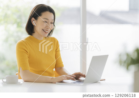 A woman in her fifties wearing earphones and facing a laptop 129206118