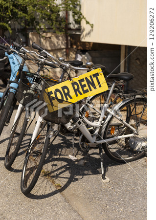 Multiple bicycles with baskets are lined up next to a wall, featuring a bright yellow sign indicating they are available for rent. The sun shines down, creating a vibrant atmosphere 129206272