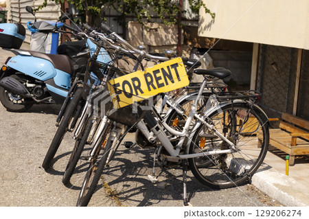 Several bicycles are parked with a prominent sign indicating they are for rent. The scene captures a vibrant outdoor environment with sunny weather and parked motorbikes nearby 129206274
