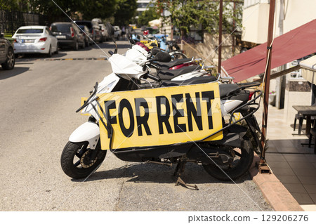 A scooter with a large yellow sign reading FOR RENT is parked along a busy street, surrounded by various cars and other parked vehicles under bright sunlight A scooter with a large yellow sign reading FOR RENT is parked along a busy street, surrounded by various cars and other parked vehicles under bright sunlight 129206276
