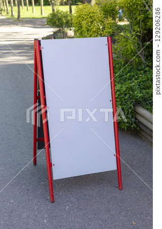 A blank outdoor sign stands on a pathway in a park filled with greenery. The sign is framed in bright red and awaits messages or advertisements. Sunlight illuminates the area. Mockup. A blank outdoor sign stands on a pathway in a park filled with greenery. The sign is framed in bright red and awaits messages or advertisements. Sunlight illuminates the area. Mockup. 129206286