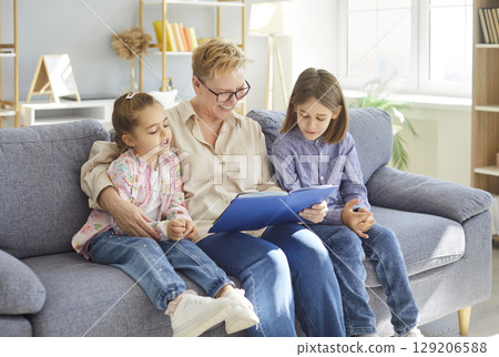 Smiling grandmother sitting on sofa and reading storybook to two children during family leisure time 129206588