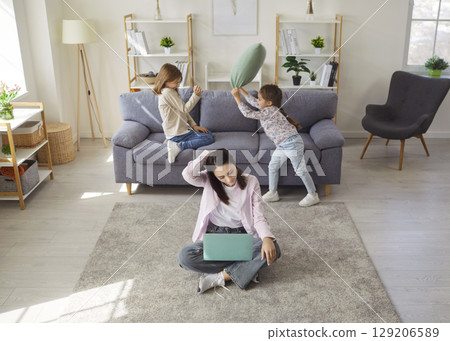 Mother working on laptop with noisy little kids playing pillow fight on sofa at home. 129206589