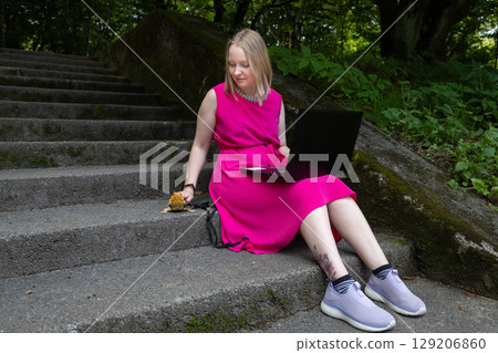 Young woman works on her laptop in a summer park, her pet agama lizard lounging peacefully beside her on a delicate leash Young woman works on her laptop in a summer park, her pet agama lizard lounging peacefully beside her on a delicate leash 129206860