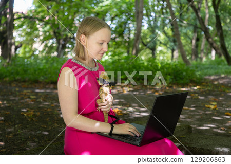 Young woman working on her laptop in a summer park gently cradles her harnessed agama lizard in a tender embrace Young woman working on her laptop in a summer park gently cradles her harnessed agama lizard in a tender embrace 129206863