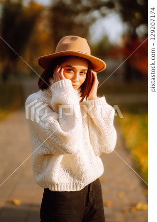 Autumn woman. Young cheerful woman in white sweater and hat walks in the nature park on a sunset. Autumn woman. Young cheerful woman in white sweater and hat walks in the nature park on a sunset. 129207174