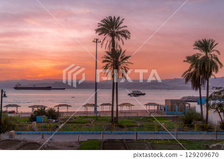Vivid sunset over Aqaba Bay, Red Sea, with silhouettes of palm trees, ships, and quiet beach huts in the resort town of Aqaba, Jordan. 129209670