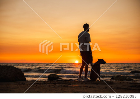 Man with a German Boxer dog sitting on a sandy beach, watching the sunset over the ocean waves. 129210083