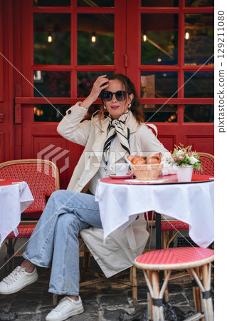 Woman in trench coat adjusts sunglasses at cafe table outdoors 129211080
