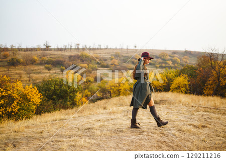 Beautiful stylish girl walks in the autumn park. The girl is dressed in a green coat and a red hat. Beautiful stylish girl walks in the autumn park. The girl is dressed in a green coat and a red hat. 129211216