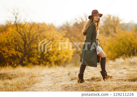 Beautiful stylish girl walks in the autumn park. The girl is dressed in a green coat and a red hat. 129211226