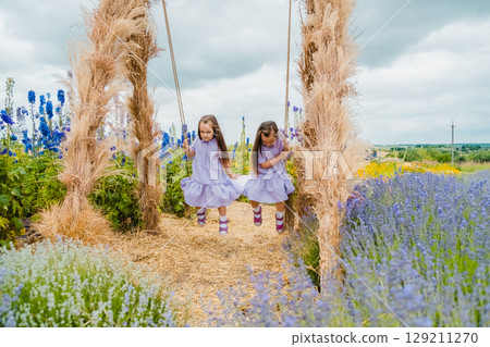 Twin girls swing on a big swing in the middle of a lavender field Twin girls swing on a big swing in the middle of a lavender field 129211270