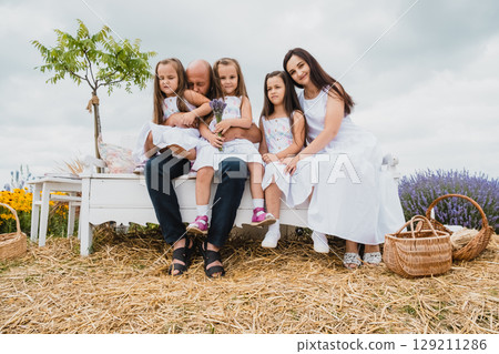 A young family sits on a white wooden retro bench in the middle of a lavender field - a father, mother and three daughters A young family sits on a white wooden retro bench in the middle of a lavender field - a father, mother and three daughters 129211286