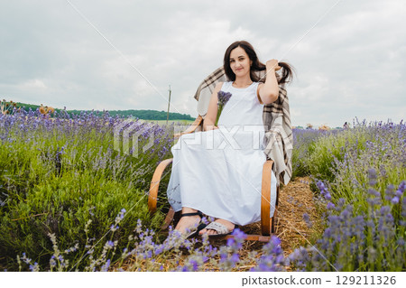 A young woman in a long white dress sits on a rocking chair in the middle of a lavender field 129211326