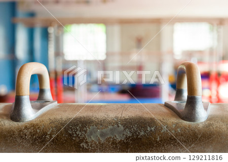 Pommel horse in gymnasium. The horse is tan leather with wooden legs and handles. Pommel horse in gymnasium. The horse is tan leather with wooden legs and handles. 129211816