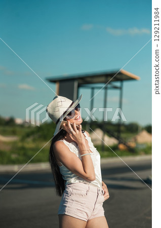 A girl wearing white shorts, a T-shirt and a hat is standing and holding the phone to her ear 129211984