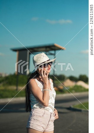 A young woman in a light white outfit is standing in the middle of nature and talking on the phone A young woman in a light white outfit is standing in the middle of nature and talking on the phone 129211985