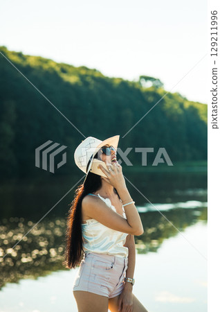 A girl in white shorts and a T-shirt stands by a lake, holding a phone to her ear 129211996