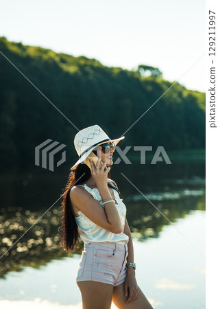 A girl in white shorts and a T-shirt stands by a lake, holding a phone to her ear 129211997