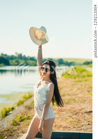 A young woman in a light-colored outfit raises her hat to the sky, as if greeting a sunny day 129212004