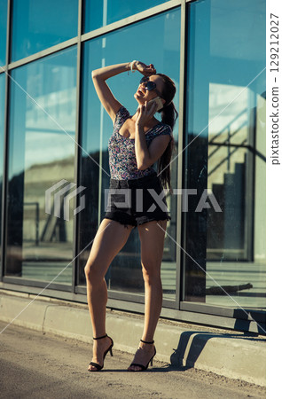 A young woman in a stylish outfit is talking on the phone next to a glass structure A young woman in a stylish outfit is talking on the phone next to a glass structure 129212027