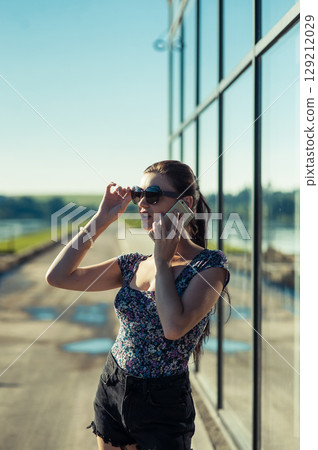 A young woman in a T-shirt and shorts is talking on the phone, standing next to a mirrored facade A young woman in a T-shirt and shorts is talking on the phone, standing next to a mirrored facade 129212029