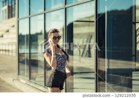 A young woman in shorts stands next to a shiny building, holding a phone A young woman in shorts stands next to a shiny building, holding a phone 129212030