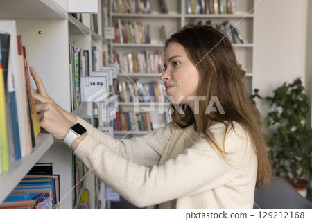 Smiling teen girl student bookstore customer select literature on bookshelf Smiling teen girl student bookstore customer select literature on bookshelf 129212168