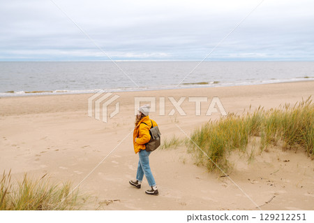Attractive woman on windy cold beach. Beautiful tourist woman in stylish clothes enjoys life by sea. 129212251