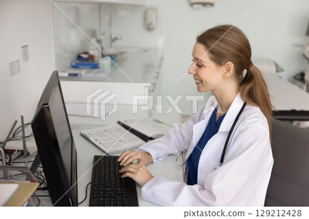 Happy young female practitioner working at desktop in doctor office Happy young female practitioner working at desktop in doctor office 129212428