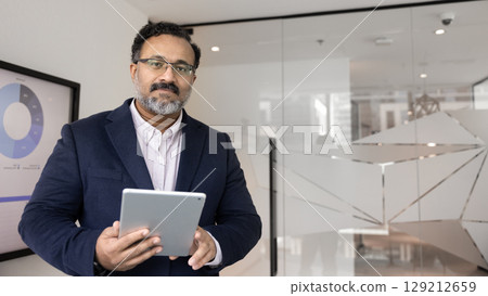 Experienced businessman in formal wear standing in boardroom holding tablet Experienced businessman in formal wear standing in boardroom holding tablet 129212659